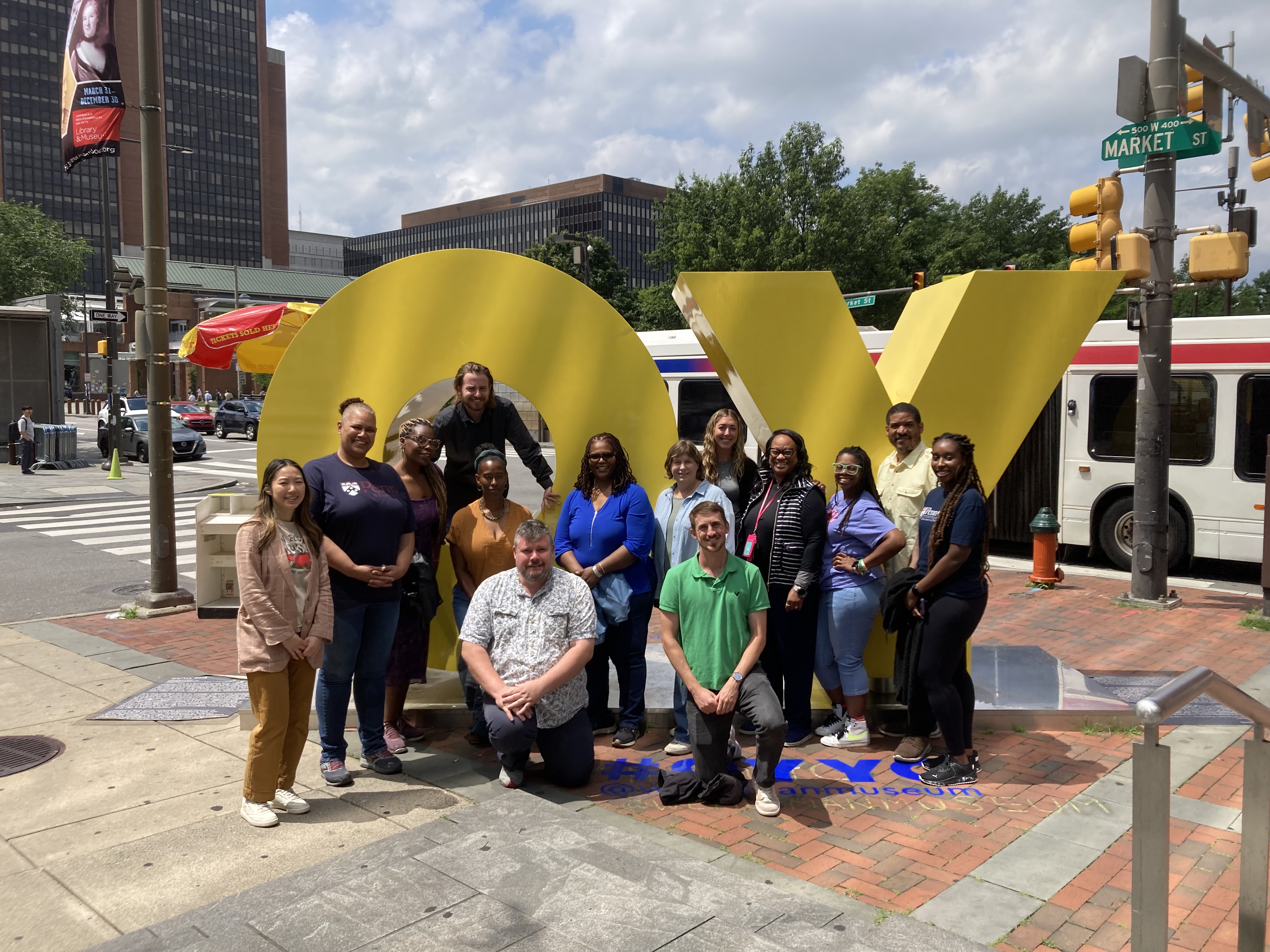 A group of adults poses outdoors in front of large, bright yellow sculptural letters at a city intersection. Some people stand while others kneel in front. Buildings, street signs, and a bus are visible in the background, indicating an urban setting. The group smiles at the camera, suggesting a team outing or organized event.