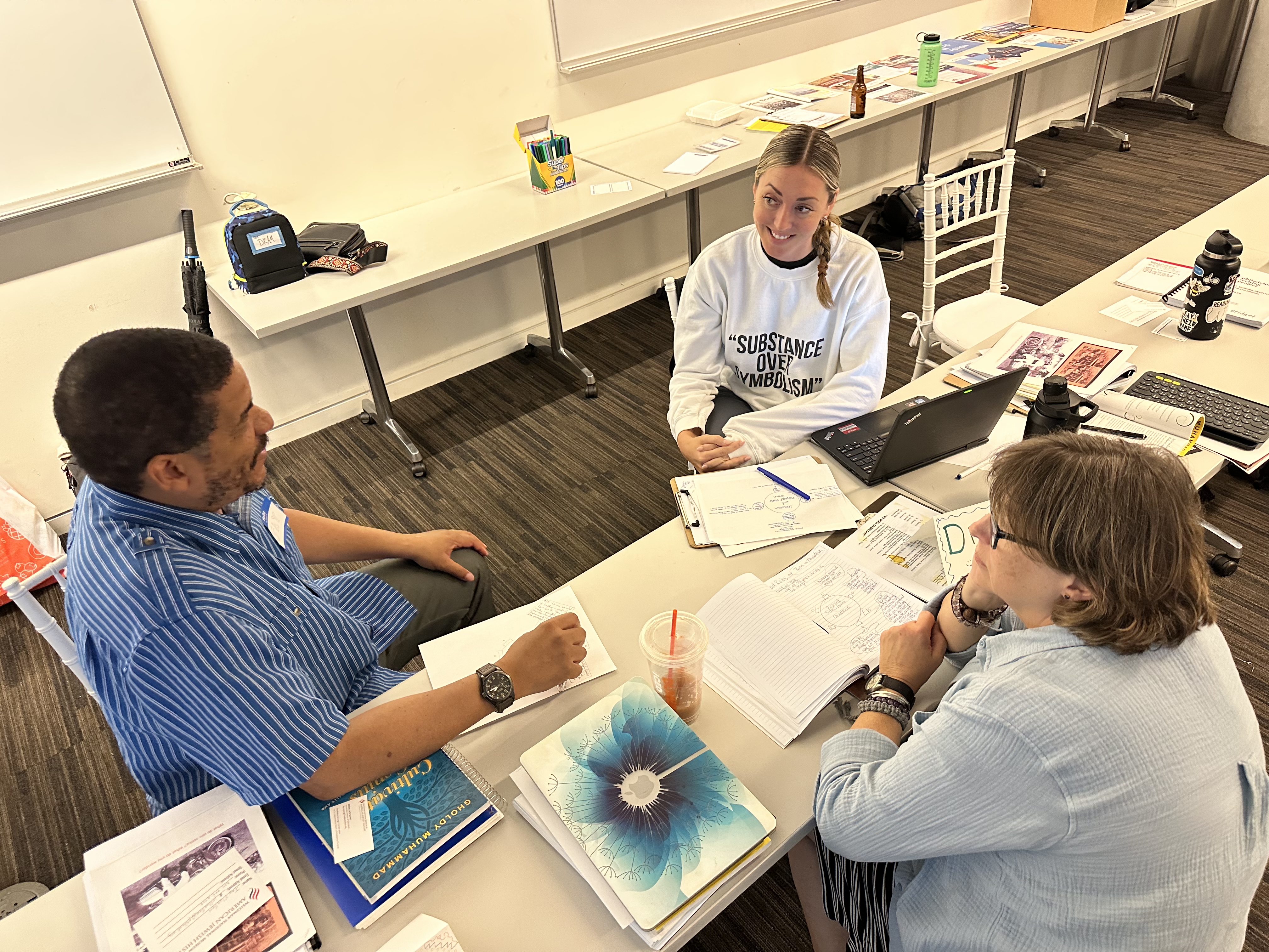 Three adults sit around a rectangular table in a classroom or workshop space, engaged in discussion. Papers, notebooks, a laptop, and books are spread across the table. One person smiles while speaking, and the others listen attentively. Whiteboards and additional materials are visible in the background, suggesting a collaborative learning or planning session.
