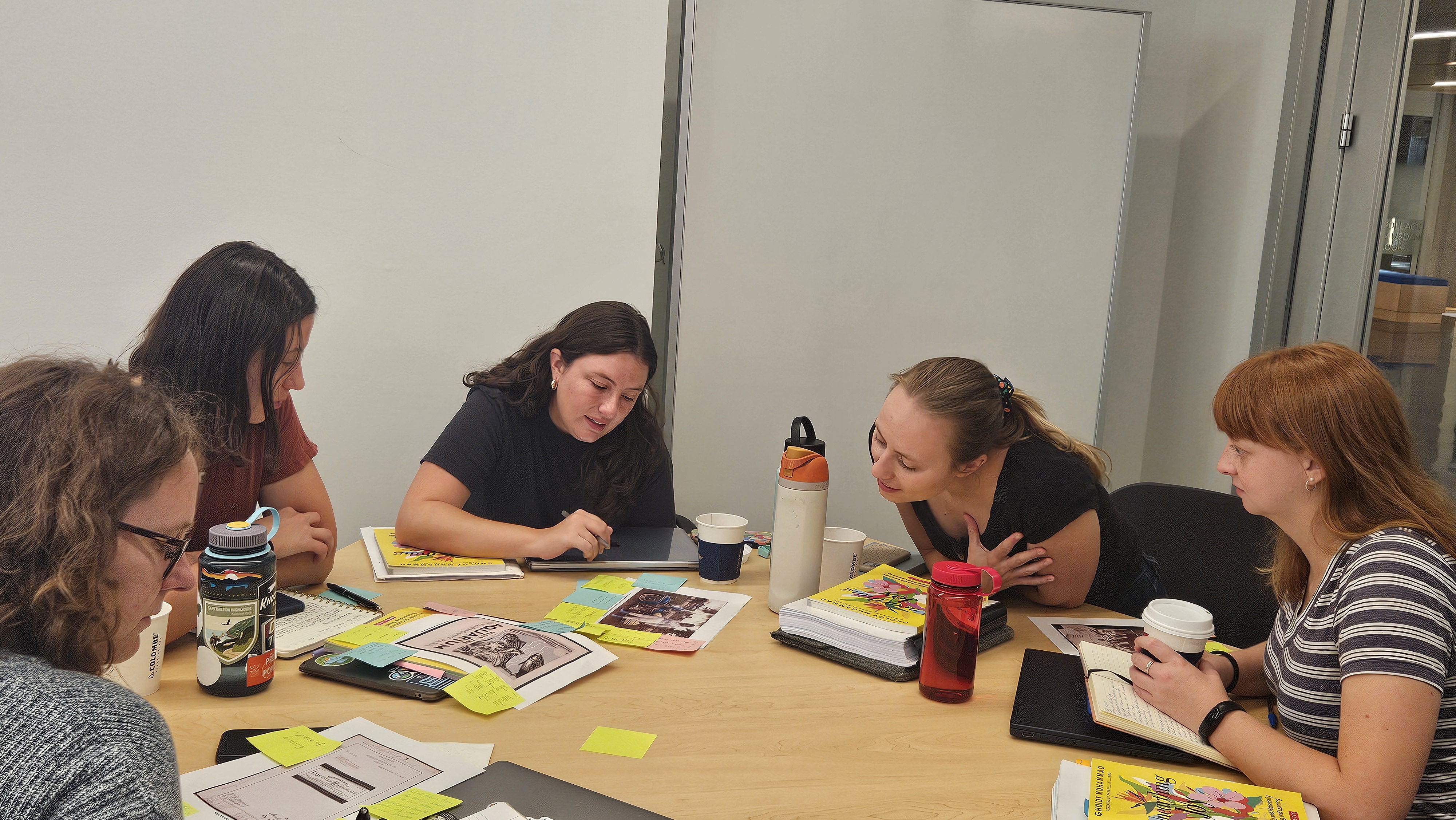 A group of four young women sitting around a circular wooden table in a collaborative meeting or study session. They are focused on various materials, including open notebooks, papers with printed images, and sticky notes. The setting appears to be a bright, modern office or classroom with white walls. Multiple water bottles and coffee cups are scattered across the table.