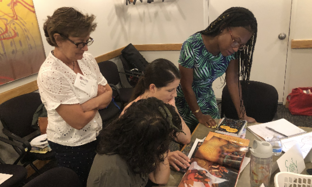 Four people are gathered around a table, looking at books and papers. Two are standing, and two are seated, engaged in a collaborative activity.