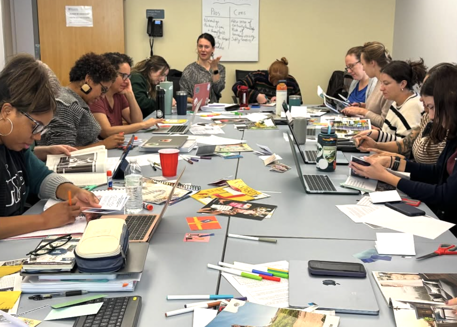 A group of approximately ten diverse women are seated around a long, grey table in a brightly lit classroom or workshop setting. They are engaged in a creative activity, with the table cluttered with open laptops, magazines, scissors, markers, and notebooks. Some participants are writing or cutting out images, while others look toward a woman at the head of the table who appears to be speaking