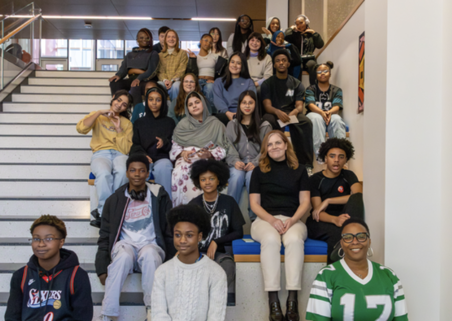 A large group of diverse young people and a few adults posing for a group photo on a wide set of modern, indoor stadium-style stairs. Most are seated on blue cushioned tiers, while others stand or sit on the steps behind them. They are dressed in a variety of casual clothing, including sweatshirts, t-shirts, and sweaters. The setting features light-colored walls and a wood-paneled ceiling with recessed lighting.