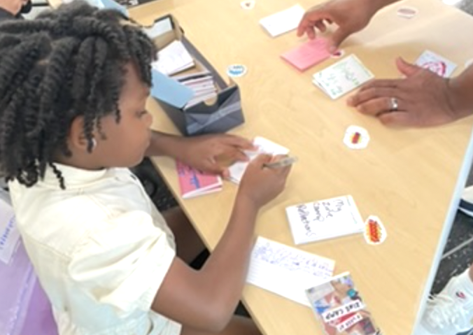 A young girl is writing a note while seated at a wooden table and is joined by an adult looking through other paper and sticky notes.